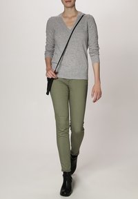 Pull en V gris, pantalon vert ajusté et bottines noires. Le mannequin pose avec un sac en bandoulière. Matériaux doux et design ajusté.