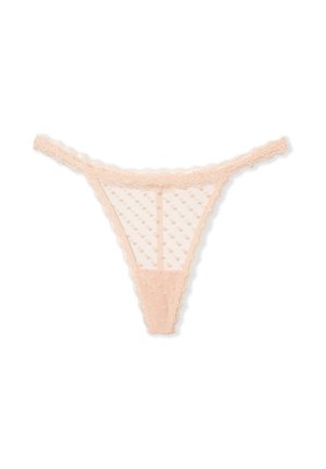 Mujer con top corto blanco sin mangas y tanga de encaje rosa transparente con puntos, de pie frente a un fondo beige.