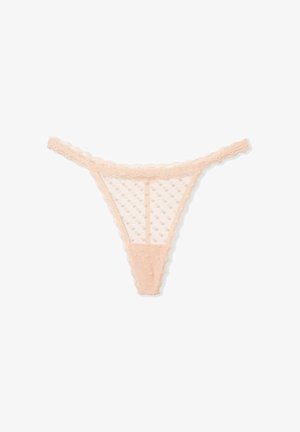 Mujer con top corto blanco sin mangas y tanga de encaje rosa transparente con puntos, de pie frente a un fondo beige.