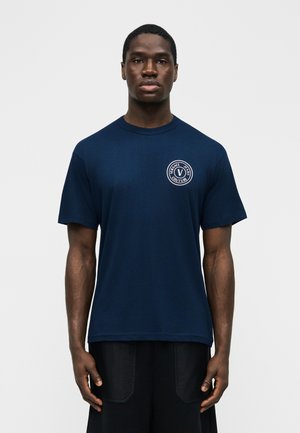Uomo che indossa una t-shirt Versace Jeans Couture blu navy con un logo circolare bianco sul petto a sinistra, in piedi davanti a uno sfondo uniforme.