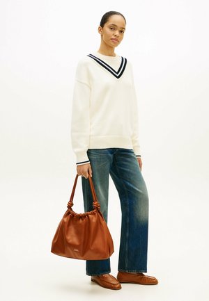 Donna che indossa un maglione bianco con scollo a V a righe nere, jeans blu, mocassini marroni, con una grande borsa di pelle marrone, in piedi su sfondo bianco.