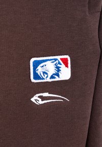 Brunt stof med et broderet mærke af en brølende vildkat i blå, hvid og rød, over et hvidt abstrakt swoosh-logo.