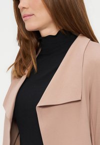 Femme aux cheveux bruns raides portant un haut noir à col montant sous un blazer rose clair, visible de la poitrine vers le haut.