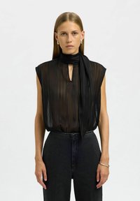 Blouse noire transparente à rayures verticales, col montant et détail d'écharpe attachée. Portée avec un jean foncé taille haute. Design minimaliste.