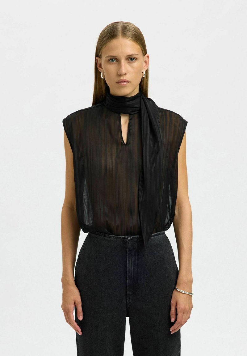 Blouse noire transparente à rayures verticales, col montant et détail d'écharpe attachée. Portée avec un jean foncé taille haute. Design minimaliste.