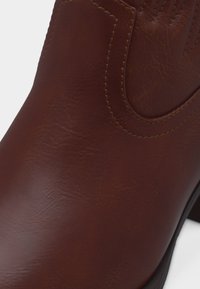 Καφέ δερμάτινη μπότα με λεπτομερείς ραφές και ελαφριά υφή, εμφανιζόμενη σε κοντινό πλάνο που εστιάζει στο μπροστινό και πλαϊνό μέρος πάνω.