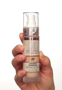 Pumpflasche mit Foundation in einem transparenten Plastikbehälter. Verfügt über eine weiße Pumpe, beiger Farbton und bedruckte Etiketten mit Marken- und Produktdetails.