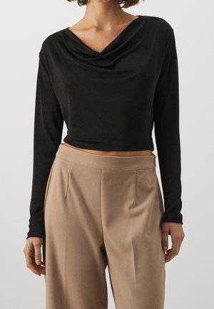 Vrouw draagt een zwarte top met lange mouwen en een gedrapeerde halslijn, gecombineerd met beige, hooggetailleerde, wijd gesneden broek, tegen een neutrale achtergrond.