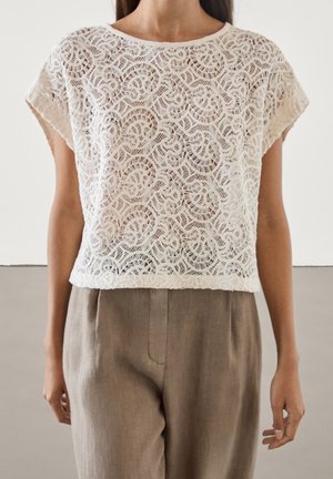 Femme portant un haut en dentelle blanche à manches courtes avec un motif circulaire et un pantalon marron taille haute, debout devant un fond uni.