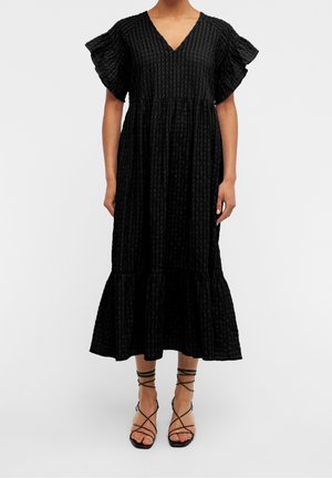 Femme portant une robe midi noire texturée avec des manches volantées et un col en V, associée à des sandales noires à talons hauts à lanières.