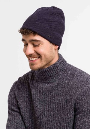 Knit navy beanie with a textured pattern and small logo accent, worn with a chunky ribbed dark gray sweater.