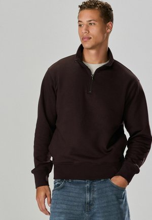 Mørkebrunt quarter-zip sweatshirt med højt krave, ribbede manchetter og kant, parret med blå jeans. Glat tekstur, afslappet design.