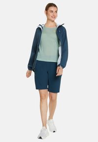 Giacca a vento navy con accenti bianchi, maglietta a manica corta verde chiaro e pantaloni corti navy. Il modello indossa scarpe da ginnastica bianche. Tessuto morbido e vestibilità casual.