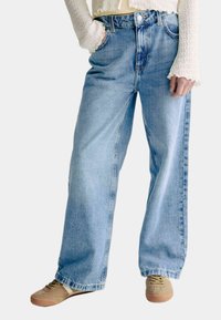 Jeans de mezclilla de pernera ancha en azul claro con una cintura alta, que cuentan con un cierre de botón y cremallera, y costuras visibles. Combinados con zapatillas marrones.