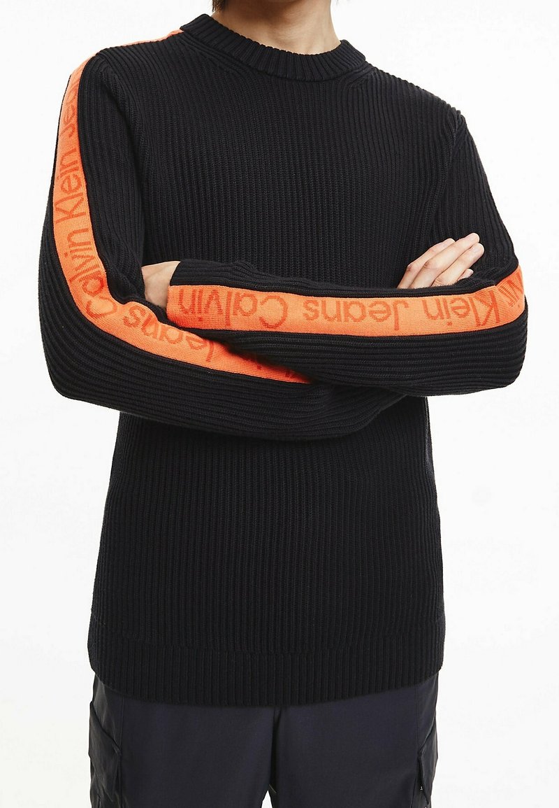 Schwarzer, gerippter Strickpullover mit orangefarbenem Calvin Klein-Logo entlang der Ärmel. Rundhalsausschnitt und eng anliegende Bündchen. Strukturierter Stoff.