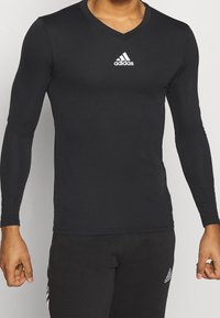 Schwarz langärmliges Sporthemd mit V-Ausschnitt, aus dehnbarem Material, mit einem weißen Adidas-Logo auf der Brust. Enganliegende Passform.