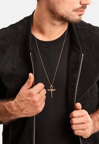 Schwarze Wildlederjacke mit Reißverschluss über einem schwarzen T-Shirt, dazu eine goldene Anhängerkette und ein gemusterter goldener Ring an der linken Hand.