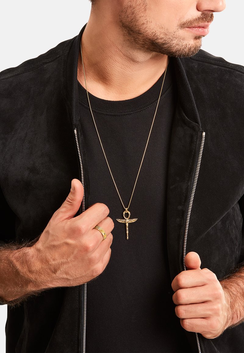 Schwarze Wildlederjacke mit Reißverschluss über einem schwarzen T-Shirt, dazu eine goldene Anhängerkette und ein gemusterter goldener Ring an der linken Hand.