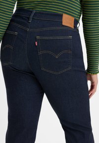 Calças de ganga azul-escuro com uma cintura alta, apresentando dois bolsos traseiros e uma etiqueta de couro com a marca na cintura. Costuras em contraste visíveis.