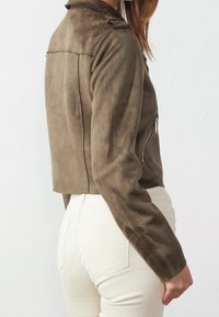 Donna che indossa un giubbotto in suede marrone corto con tasca con zip e pantaloni bianchi a vita alta, in piedi davanti a uno sfondo bianco semplice.