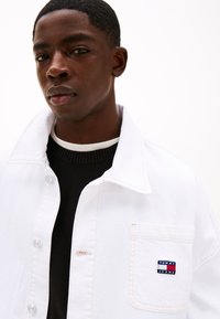 Balta džinsinė striukė su sulankstomu apykakle, priekinėmis kišenėmis ir mažesniu siuvinėtu „TOMMY JEANS“ logo. Juodas megztinis matomas po ja.