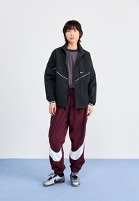 Svart vindjakke med hvite detaljer, stripete skjorte, maroon harem bukser med hvit swoosh-design, og metallisk grå joggesko.