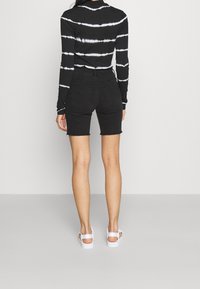 Persona que lleva pantalones cortos de mezclilla de tiro alto negros y una blusa negra de manga larga con rayas horizontales blancas, de espaldas, sobre un fondo neutro.