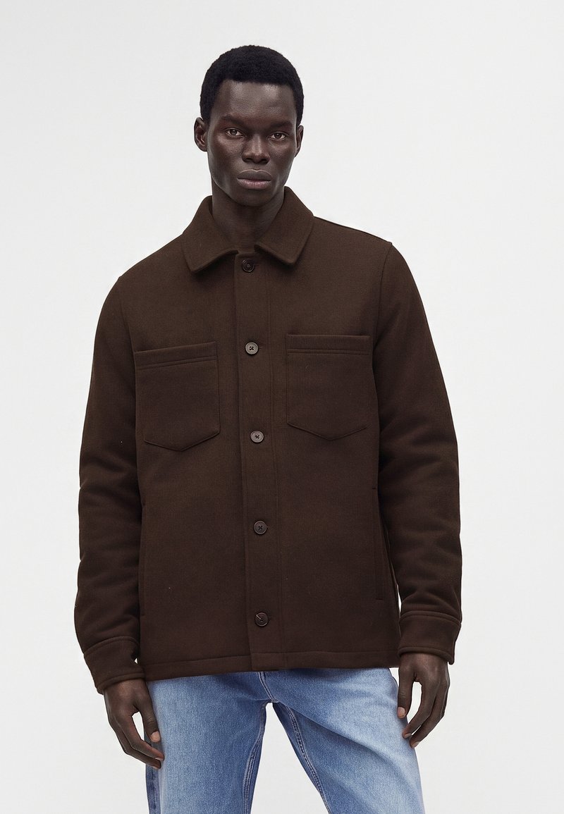 Jeune homme à la peau foncée portant une veste marron foncé boutonnée et un jean bleu clair, debout devant un fond blanc uni.