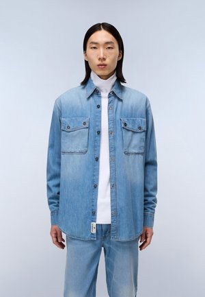 Lichtblauw denim overhemd met knoopzakken op de borst, gedragen over een witte coltrui. Het heeft een puntkraag en een rechte zoom.