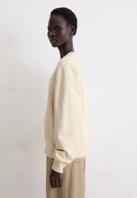 Crème-kleurige sweatshirt met een ontspannen pasvorm, ribgebreide manchetten en een ronde halslijn, gedragen met beige broeken. Minimale stikseldetails zichtbaar.