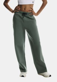 Green, smooth fabric sweatpants with a relaxed fit, drawstring waist, and side slits, paired with white athletic shoes.