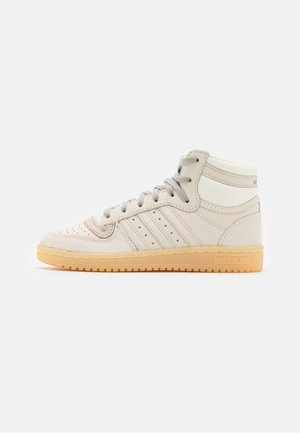 Beżowy sneaker adidas z wysoką cholewką, perforacjami, trzema paskami, szarymi sznurówkami i gumową podeszwą, widoczny z boku na białym tle.