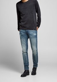 Chemise noire à manches longues avec un col rond, associée à un jean bleu clair avec des détails déchirés et des bottes noires à lacets.