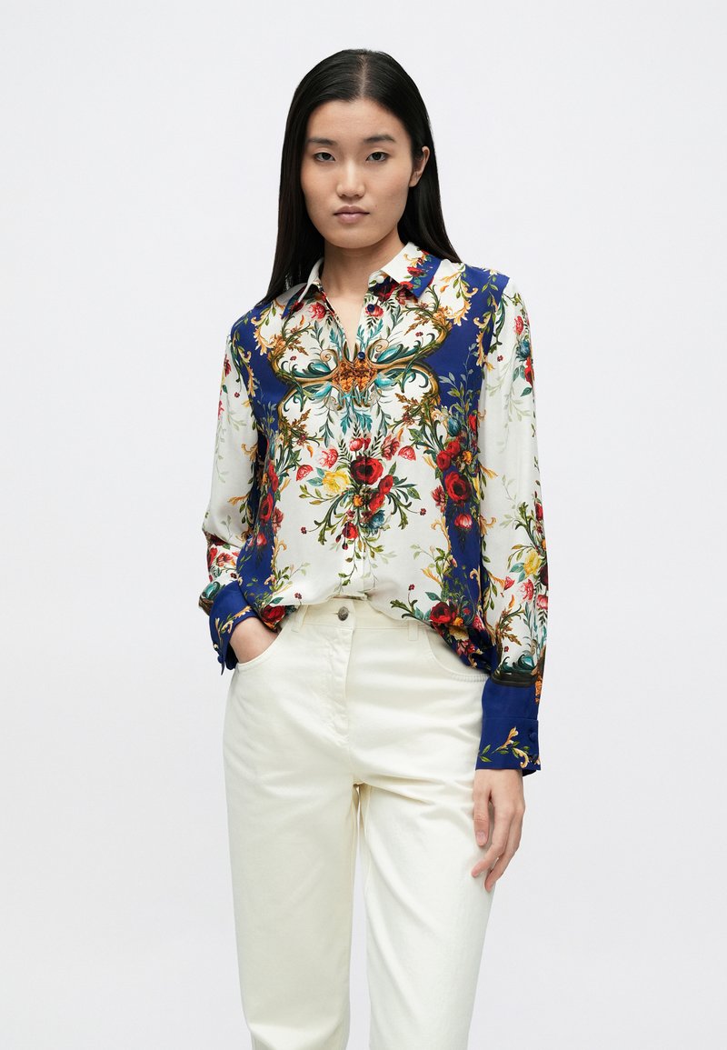 Blúzka na gombíky s kvetinovým vzorom v žiarivých farbách na krémovom pozadí, s modrými akcentmi na golieri a manžetách.