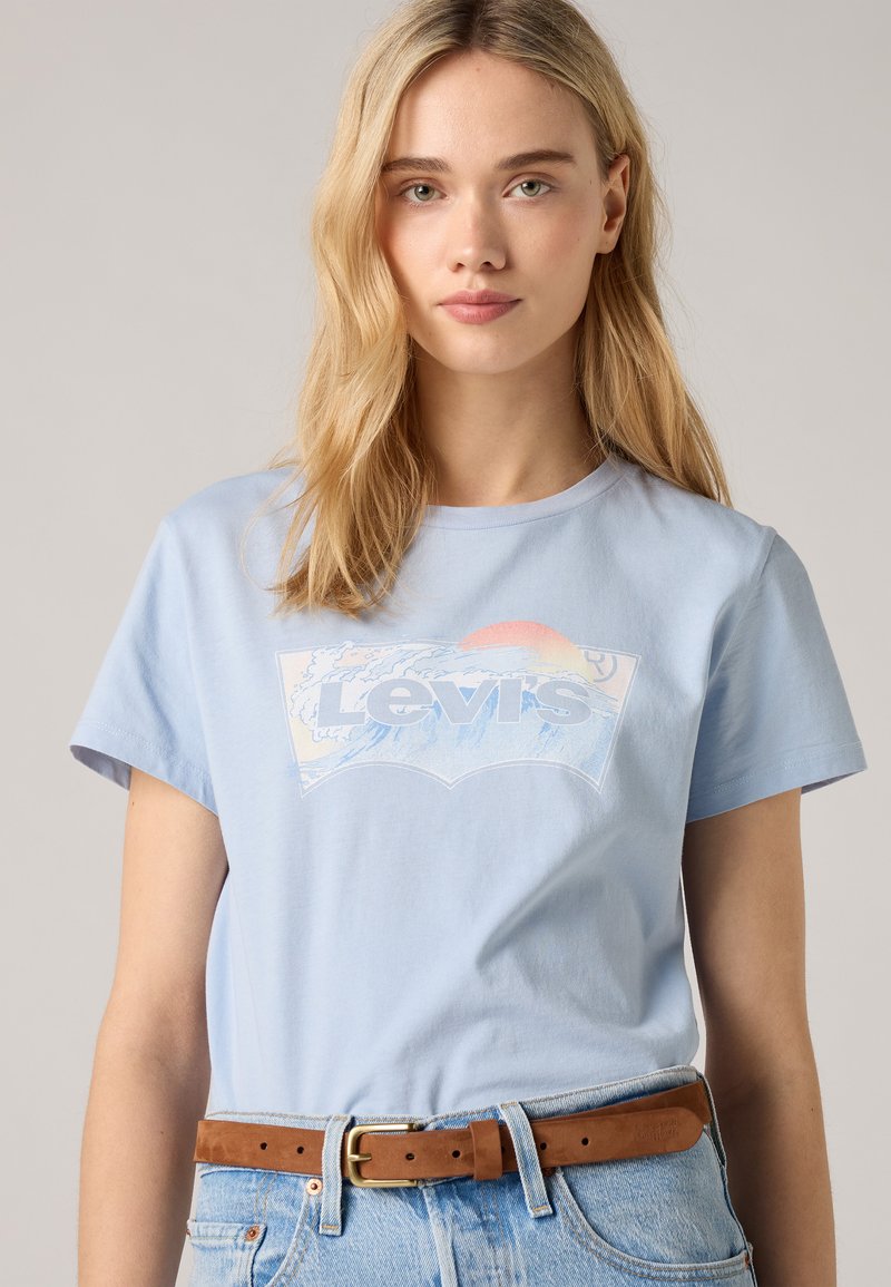 Camiseta de algodón de color azul claro con un gráfico de ola y sol con el texto "Levi's" en negrita. Mangas cortas y un corte relajado. Lleva un cinturón marrón.