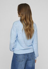 Šviesiai mėlynas megztinis su perforuotu raštu, laisvo kirpimo, apvaliu iškirpimu ir raštuotais rankogaliais. Derinamas su tamsiais džinsais.