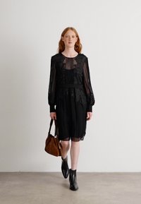 Robe noire avec des manches longues transparentes et des détails floraux brodés ; ceinture à la taille. Le modèle tient un sac marron et porte des bottines noires.