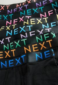 Ropa interior negra con diversas cinturas elásticas, cada una con la palabra "NEXT" en diferentes colores pasteles: rosa, morado, verde, azul y naranja.