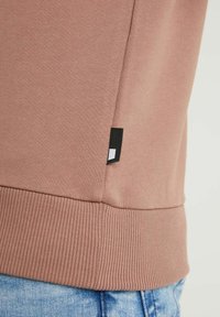 Hnědá mikina se žebrovaným lemem a bočními švy. Obsahuje malou černou nášivku blízko spodního okraje. Měkká látka a volný design.