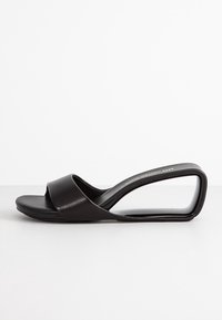 Infradito neri con due ampie fasce, suola piatta e un design unico del tacco curvo. Realizzate in pelle liscia con una finitura opaca.