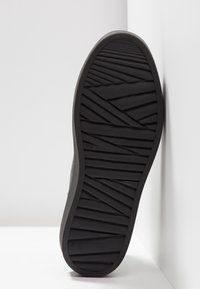 Semelle de chaussure noire avec motifs de bande de roulement diagonaux et horizontaux, présentée verticalement contre une surface d'angle blanche.