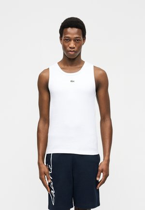 Jeune homme portant un débardeur blanc avec un petit logo et un short bleu marine avec une inscription blanche sur le côté, debout devant un fond uni.