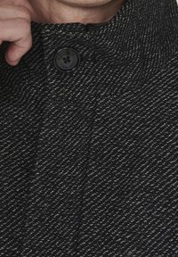 Kołnierz płaszcza w czarno-białym wzorze przypominającym rybi szew, z okrągłym czarnym guzikiem i widoczną fakturą materiału, podkreślający dopasowany fason.