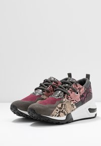 Un paio di sneakers multicolori con stampa di serpente, suede bordeaux e grigio, lacci neri e suole bianche spesse, su una superficie bianca e uno sfondo grigio chiaro.