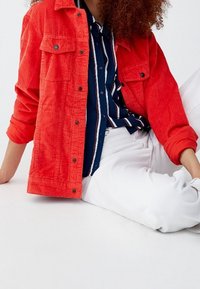 Veste en velours côtelé rouge avec deux poches poitrine, associée à une chemise rayée marine et un pantalon blanc. La veste comporte des boutons et des manches retroussées.