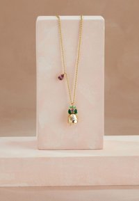 Guldhalsband med ett hänge format som en detaljerad figur prydd med gröna och lila ädelstenar, visas på ett mjukt rosa ställ.