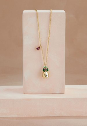 Zlatý náhrdelník s přívěskem ve tvaru detailní postavy zdobené zelenými a fialovými drahokamy, vystavený na měkkém růžovém stojanu.