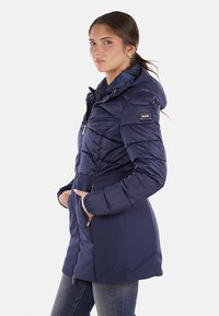 Cappotto imbottito blu navy con cappuccio, design a righe orizzontali, chiusura con zip e tasche laterali. Presenta una patch con logo sulla manica.