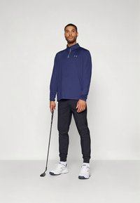 Laivastonsininen pitkähihainen pusero, jossa on vetoketjullinen kaulus, yhdistetty mustiin kapeneviin housuihin. Valkoiset urheilukengät, joissa sinisiä yksityiskohtia, ja kädessä golfmaila.