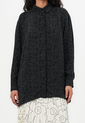 Femme portant une chemise noire à manches longues avec des contours floraux foncés sur une jupe blanche avec de grands imprimés floraux noirs.